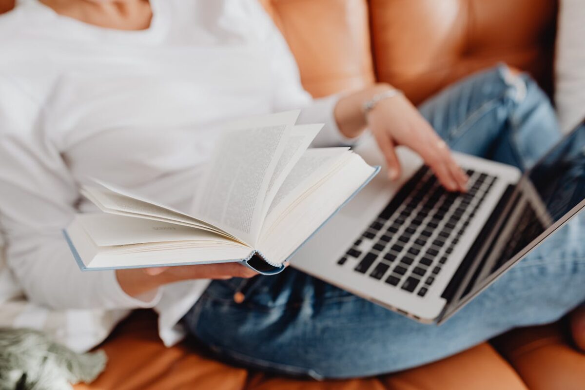 Woman sitting on a sofa, reading a book while using a laptop, symbolizing leisure and multitasking.